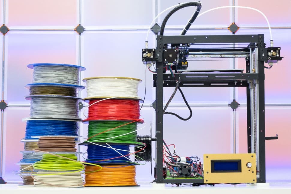 Stacked spools of colored filament beside an open frame 3D printer