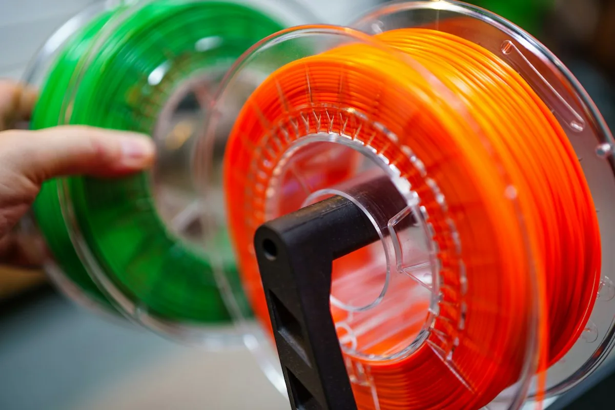 Two spools of 3D printer filament, orange and green, on a spool holder